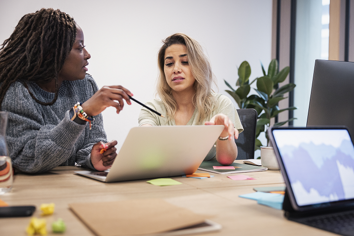 Två kvinnor sitter vid ett skrivbord med datorer och diskuterar en affärsidé.